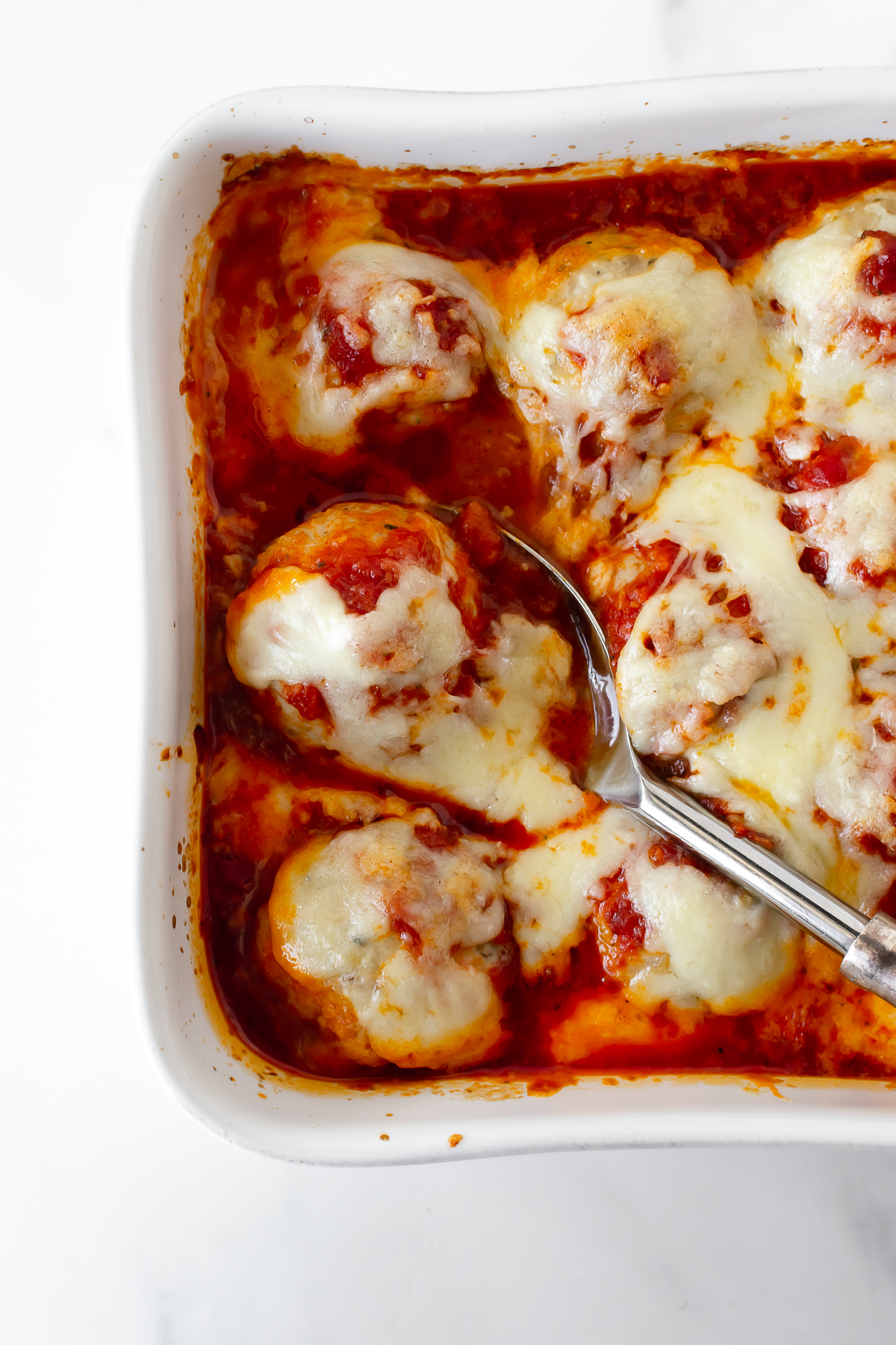 chicken parmesan meatballs in a white baking dish with marinara sauce and melted cheese with a spoon.