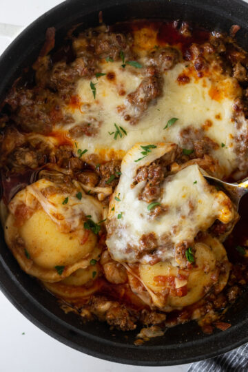 baked ravioli in a black pot with melted cheese and a serving spoon with a blue napkin.