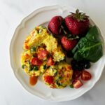 egg cups on a white plate with hot sauce, strawberries and spinach.