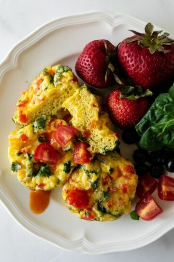 egg cups on a white plate with hot sauce, strawberries and spinach.