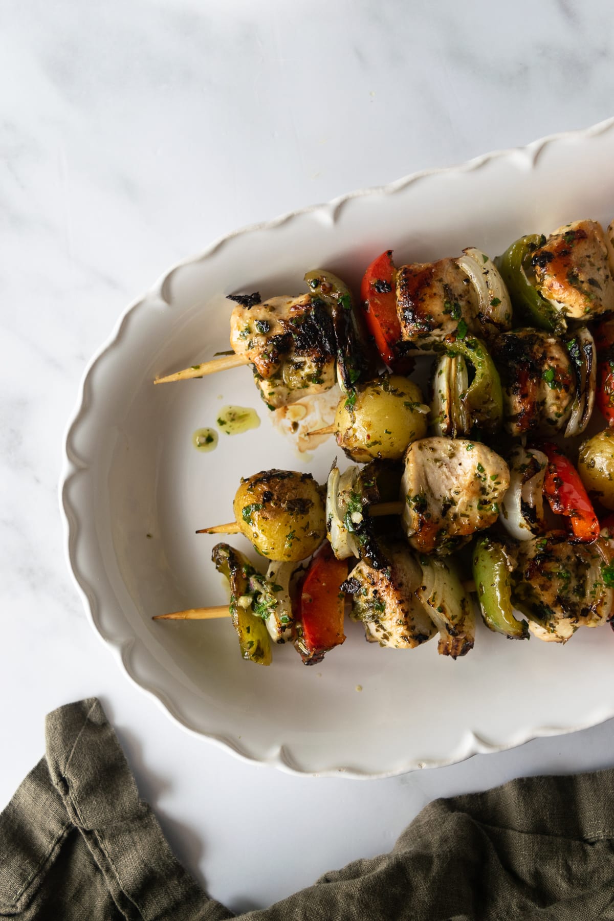 cooked chicken chimichurri on skewers with potatoes and vegetables on a white platter.