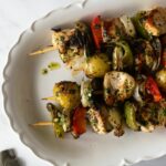 cooked chimichurri chicken on skewers with potatoes and vegetables on a white platter.