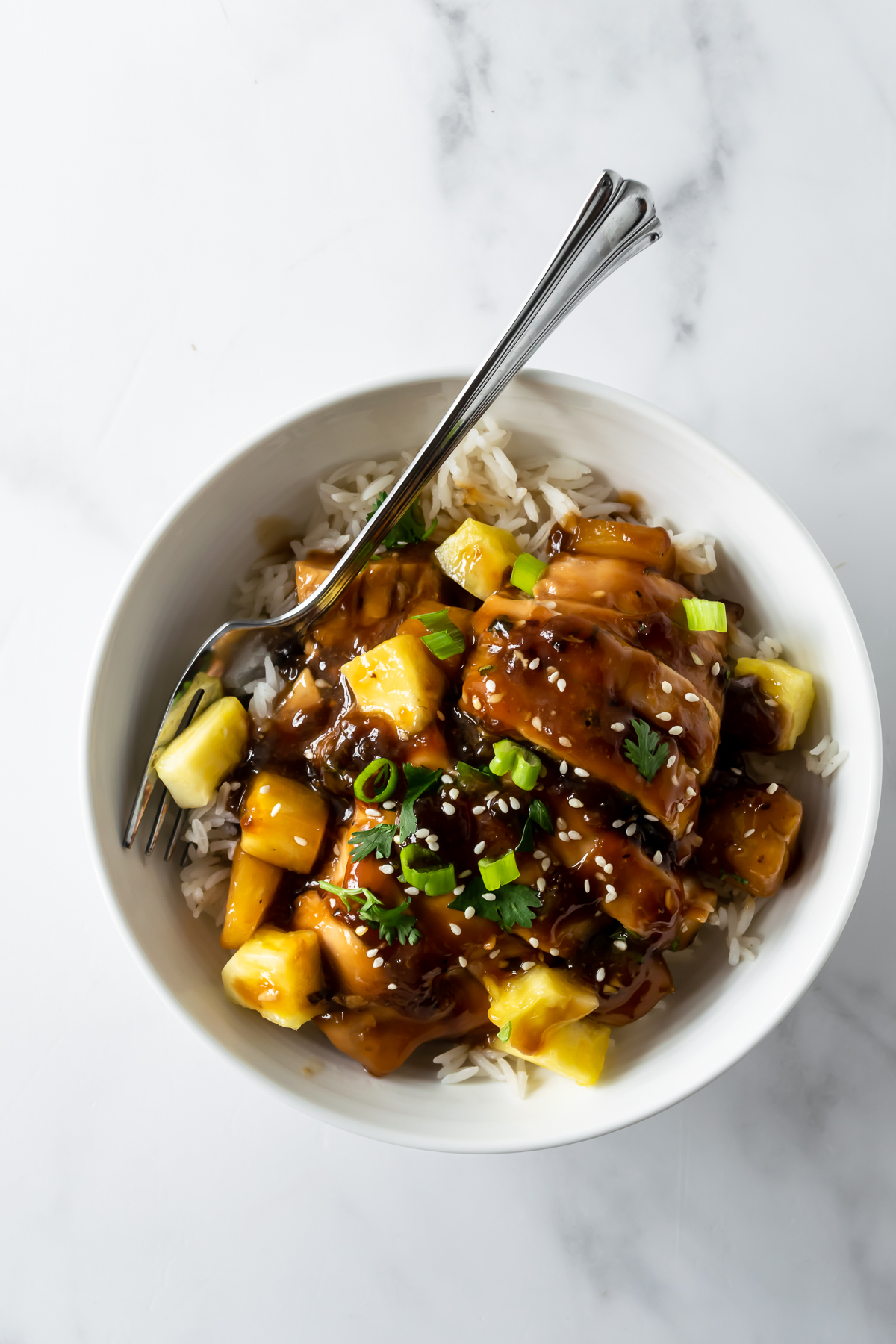 teriyaki pineapple chicken in a white bowl with a fork on rice and topped with sesame seeds.
