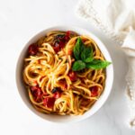 burst cherry tomato pasta in a white bowl with tomatoes and basil on a white table with a napkin.