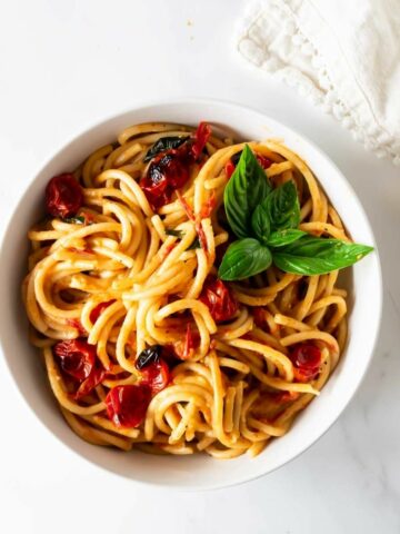 burst cherry tomato pasta in a white bowl with tomatoes and basil on a white table with a napkin.