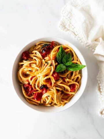 burst cherry tomatoes with pasta in a bowl topped with tomatoes and basil on a white table with a napkin.