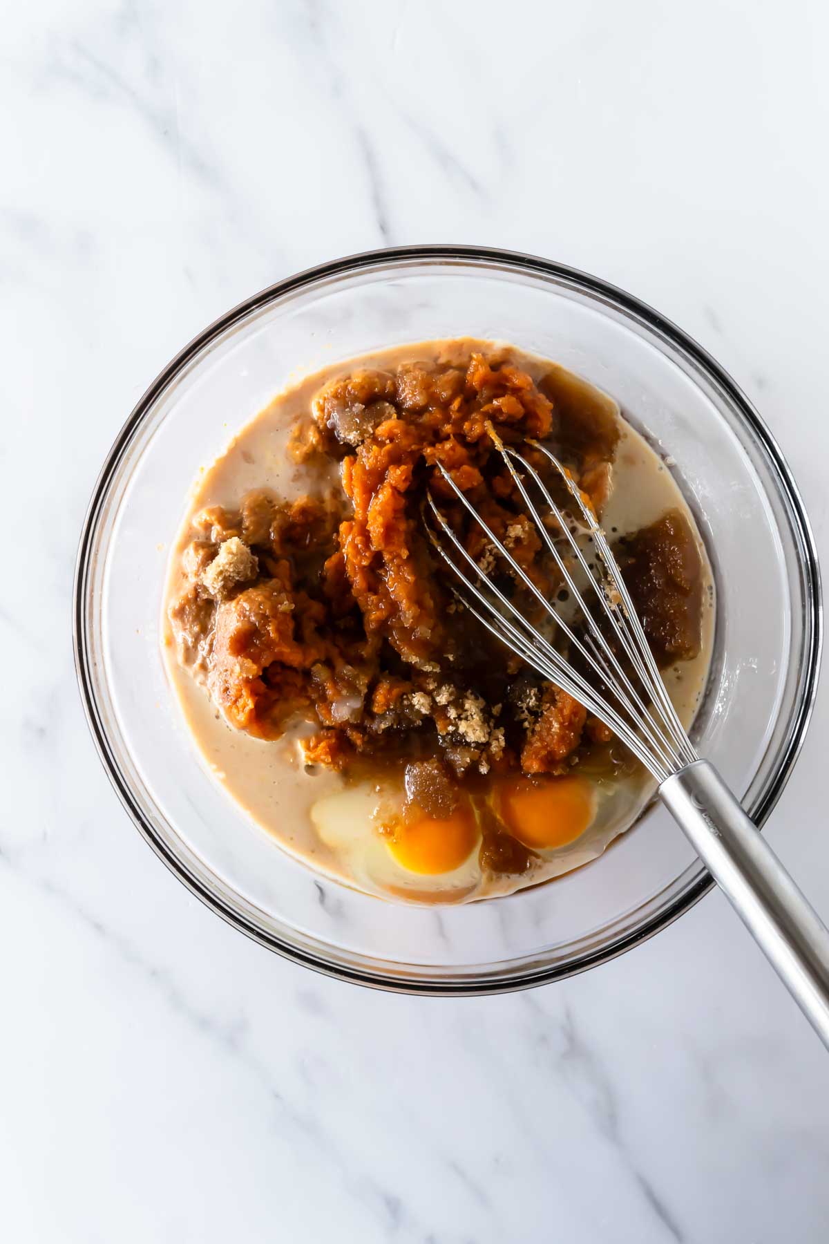 wet ingredients like pumpkin puree, eggs, sugar and vanilla in a mixing bowl with a whisk on a white table.