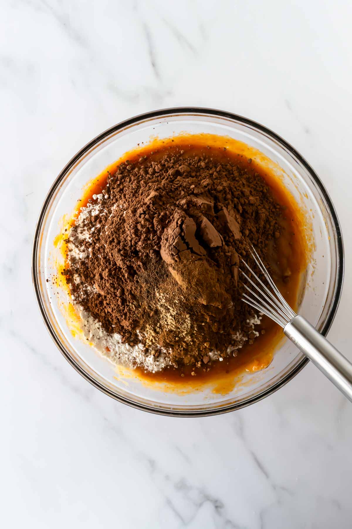 flour and cocoa powder on top of wet ingredients a mixing bowl with a whisk on a white table.