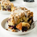 slice of blueberry crumb cake with vanilla icing on a white plate with a fork.