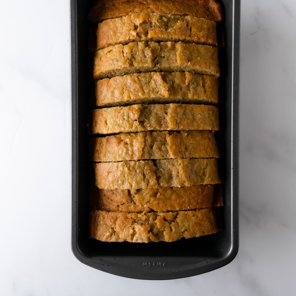 zucchini bread cut into slices in a loaf pan on a white table.