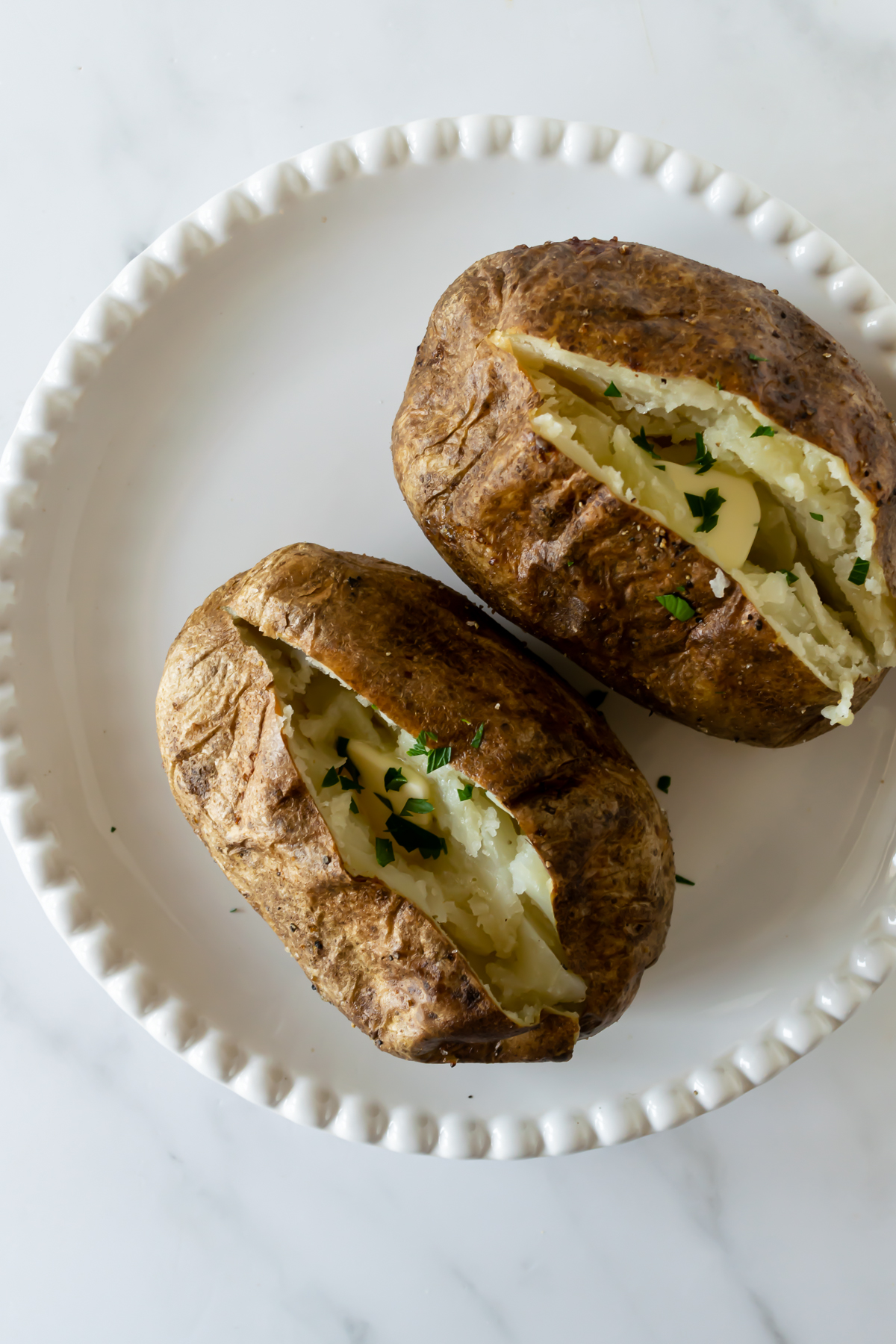 two baked potatoes on a white plate sliced open with chopped parsley.