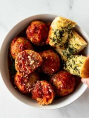 crockpot meatballs in a white bowl with a hand grabbing garlic bread.