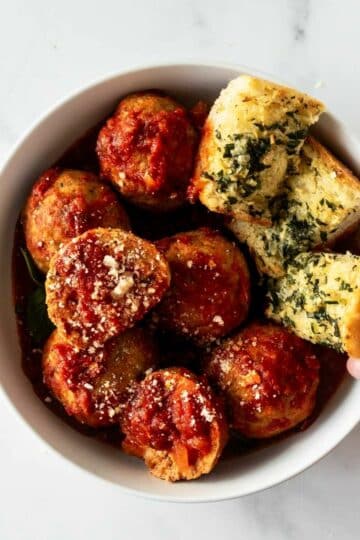 crockpot meatballs in a white bowl with a hand grabbing garlic bread.