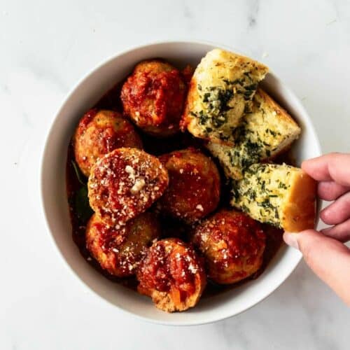 crockpot meatballs in a white bowl with a hand grabbing garlic bread.