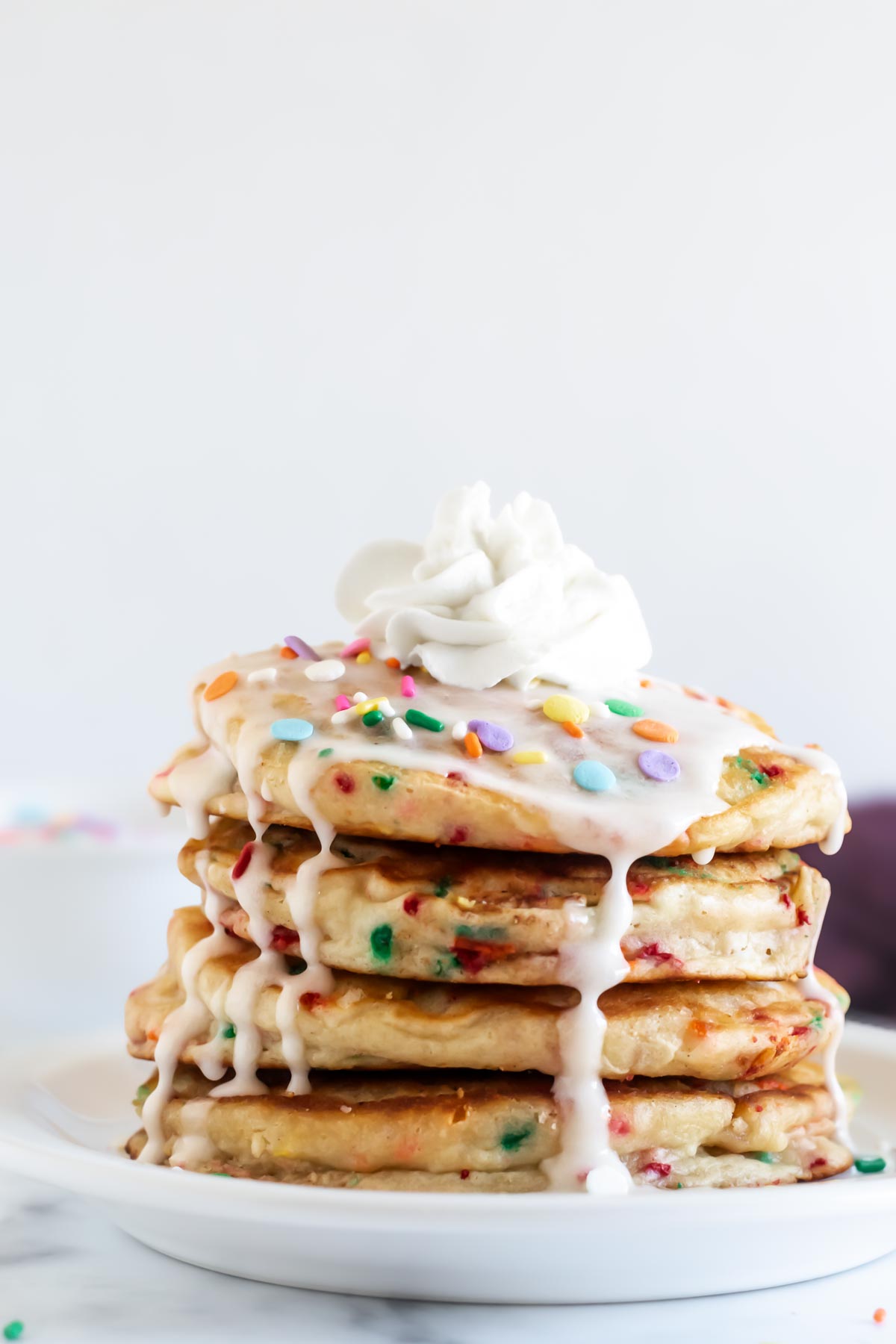 stack of funfetti birthday pancakes on a white plate with vanilla icing, sprinkles and whipped cream on top.