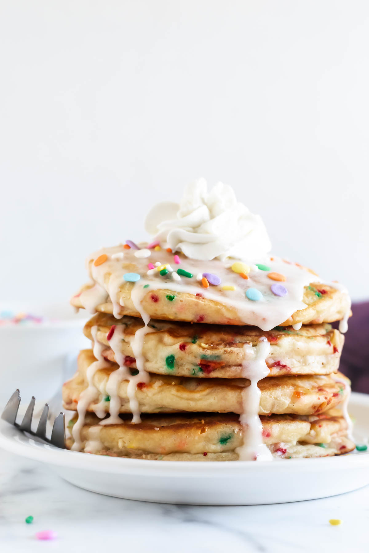 a stack of birthday cake pancakes topped with vanilla glaze, sprinkles, and whipped cream with a fork.