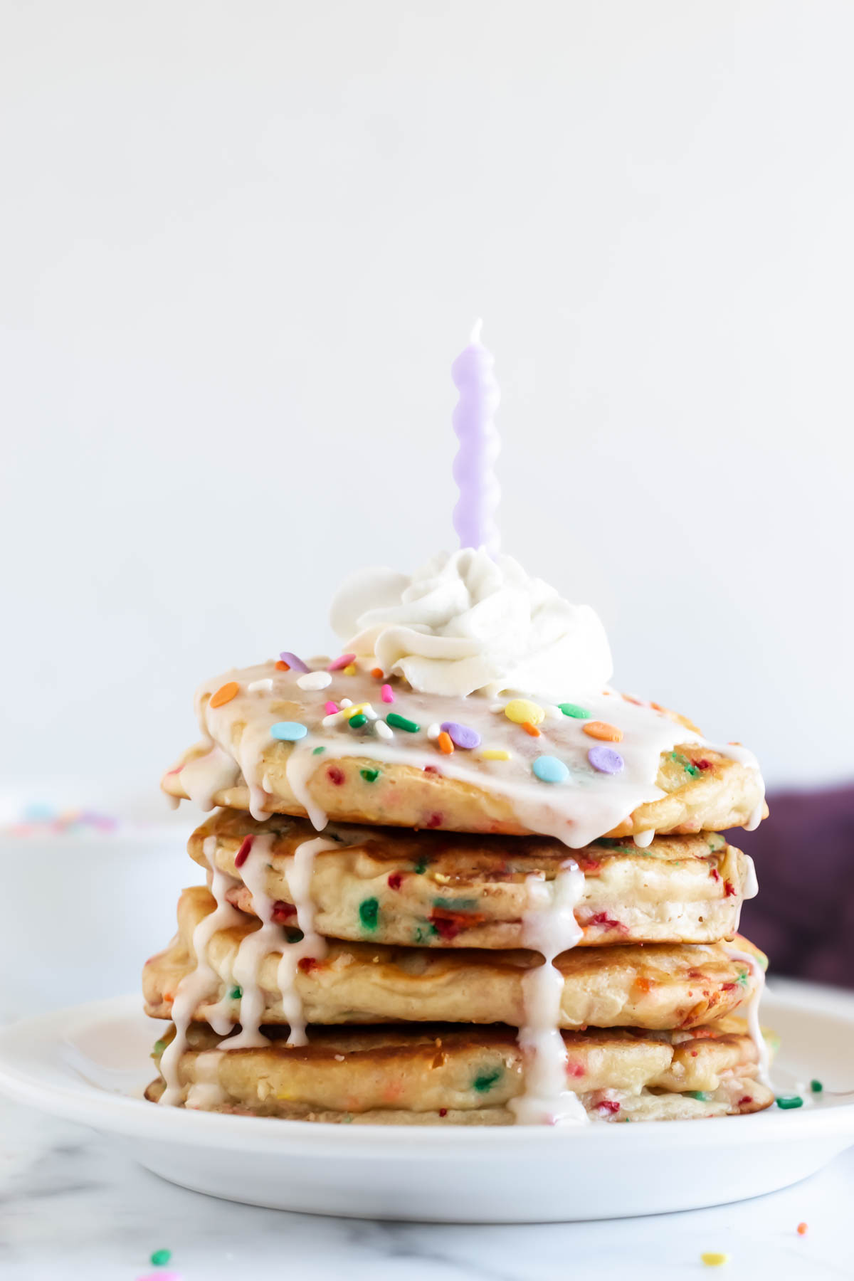 a stack of birthday cake pancakes topped with vanilla glaze, sprinkles, whipped cream, and a candle.