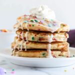 four pancakes stacked on a white plate topped with whipped cream, vanilla icing and sprinkles.