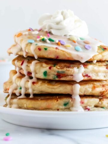 four pancakes stacked on a white plate topped with whipped cream, vanilla icing and sprinkles.