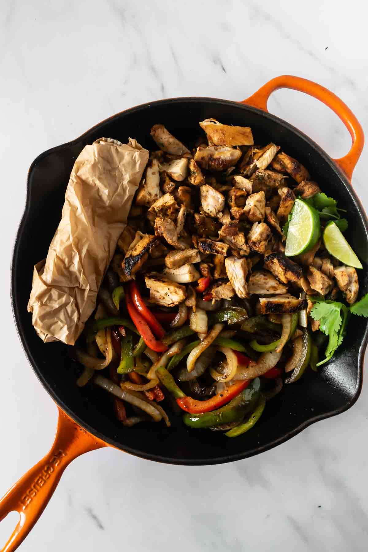 chicken fajitas in a cast iron skillet with lime slices and tortillas wrapped in parchment paper.