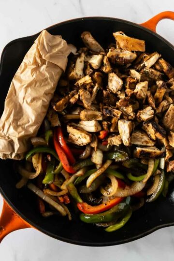 chicken fajitas in a black and orange skillet with tortillas rolled in parchment paper.