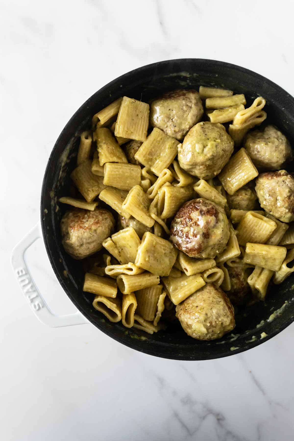 pesto and heavy cream sauce mixed with rigatoni and meatballs in a black pot.