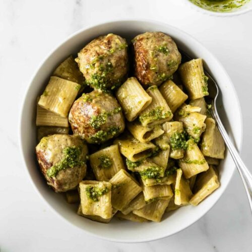 chicken pesto meatballs with pasta topped with basil pesto in a white bowl with a fork.