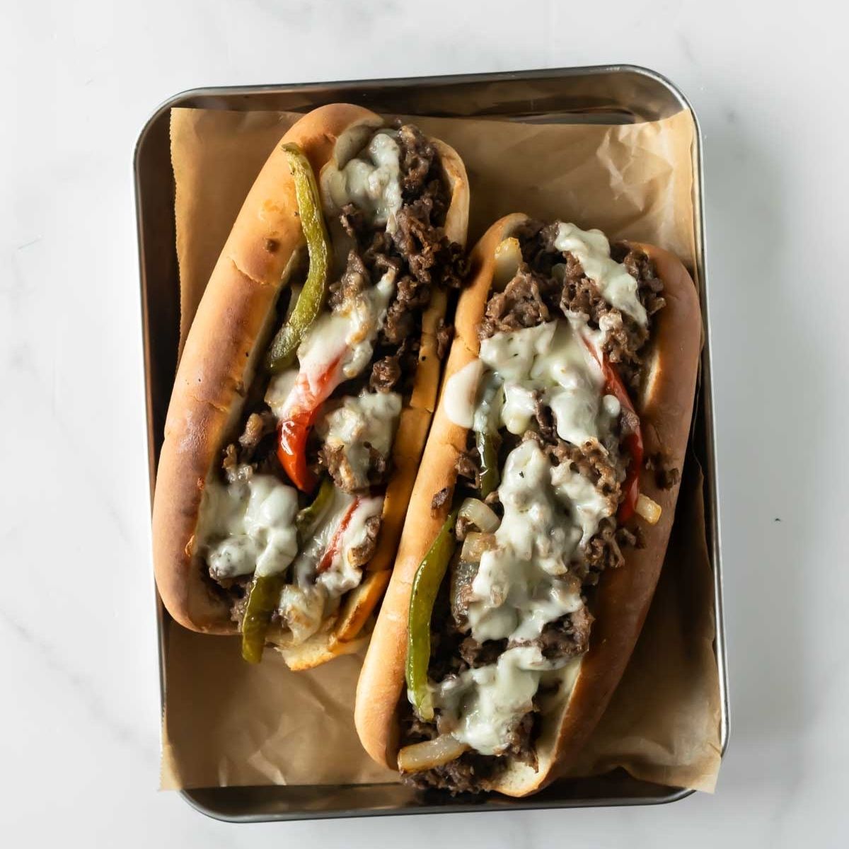 two homemade philly cheesesteaks on a tray with parchment paper topped with melted cheese.