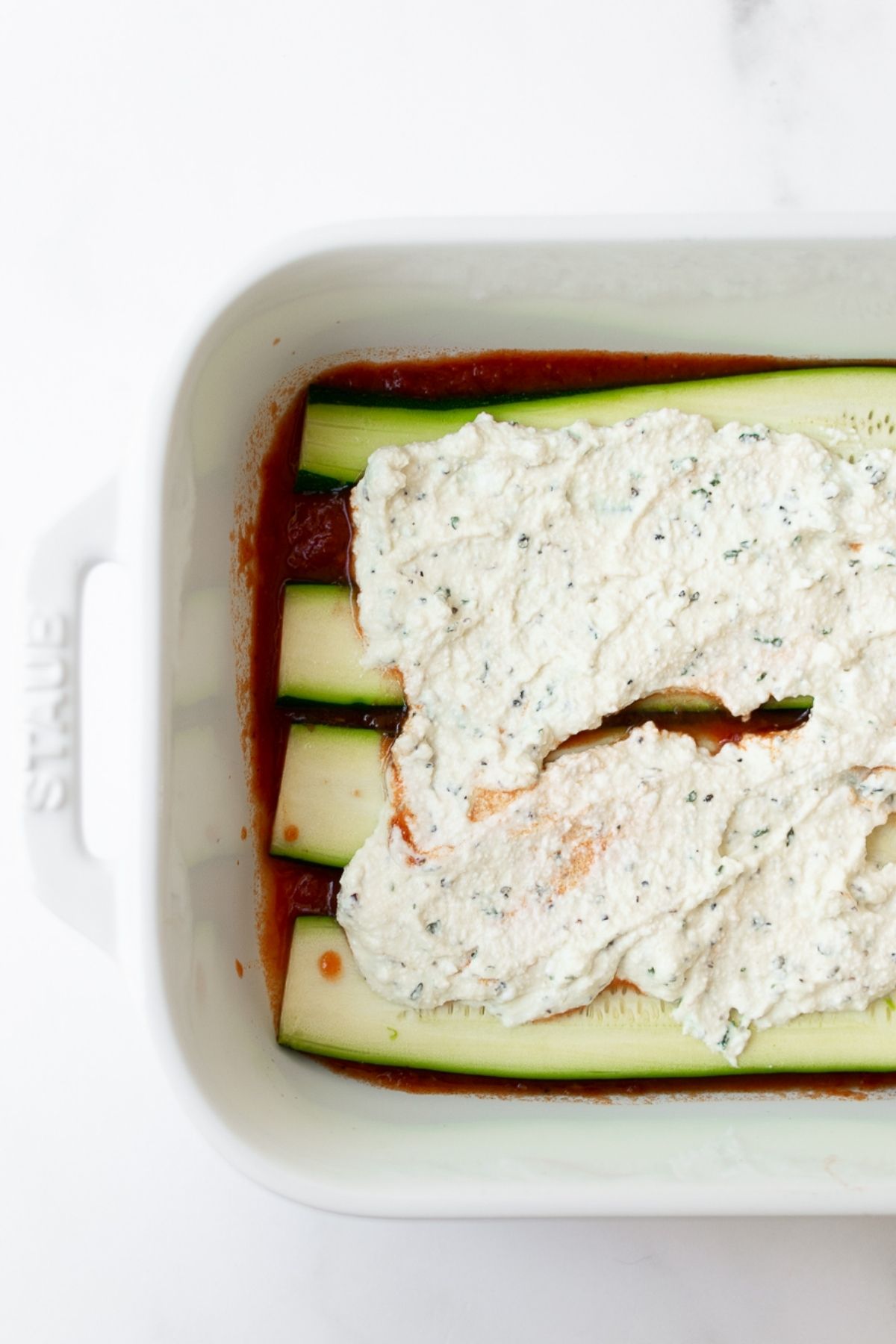 zucchini slices layered in a white baking dish topped with herbed ricotta cheese.