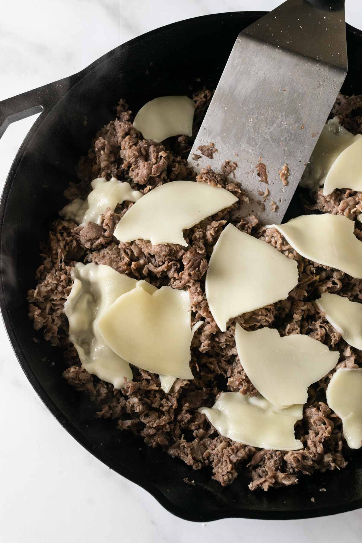 cooked shaved beef topped with slices of cheese in a cast iron skillet with a metal spatula.