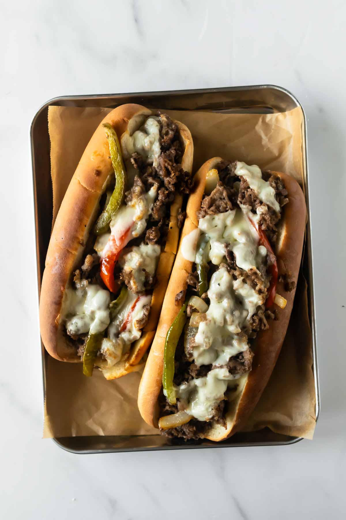 two Philly cheesesteaks sitting side by side on a tray and topped with peppers, onions, and melted cheese.