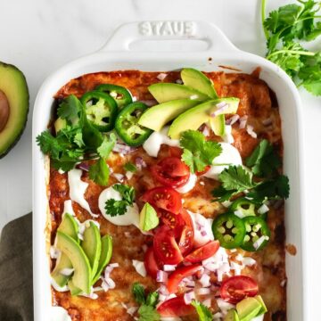 chicken enchiladas in a white baking pan topped with avocados, tomatoes and sour cream.