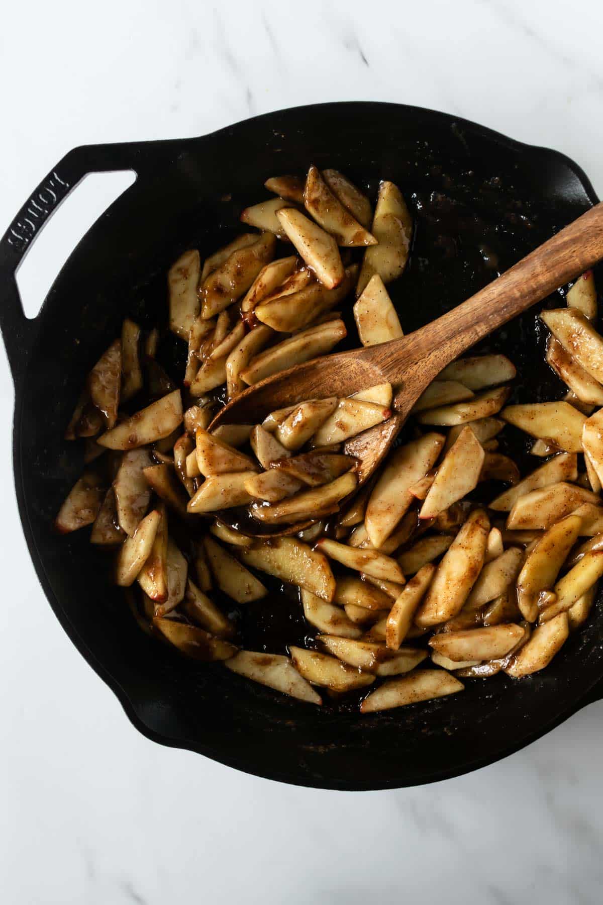 cinnamon apples in a cast iron skillet with brown sugar, spices and a wooden spoon.