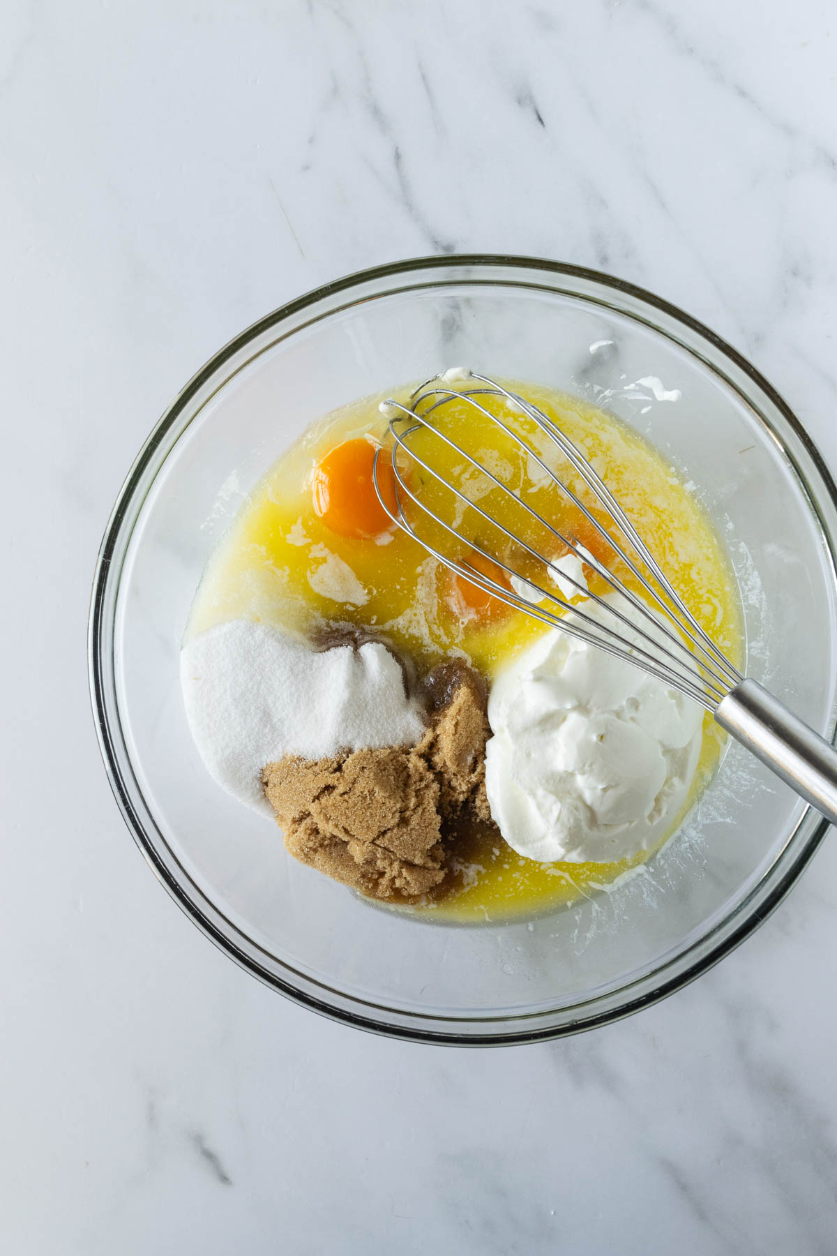 eggs, butter, sugars, and yogurt with a whisk in a clear bowl on a white table with a whisk.