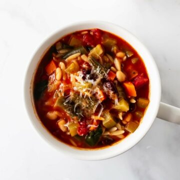 minestrone soup in a white bowl with a handle on a white table.