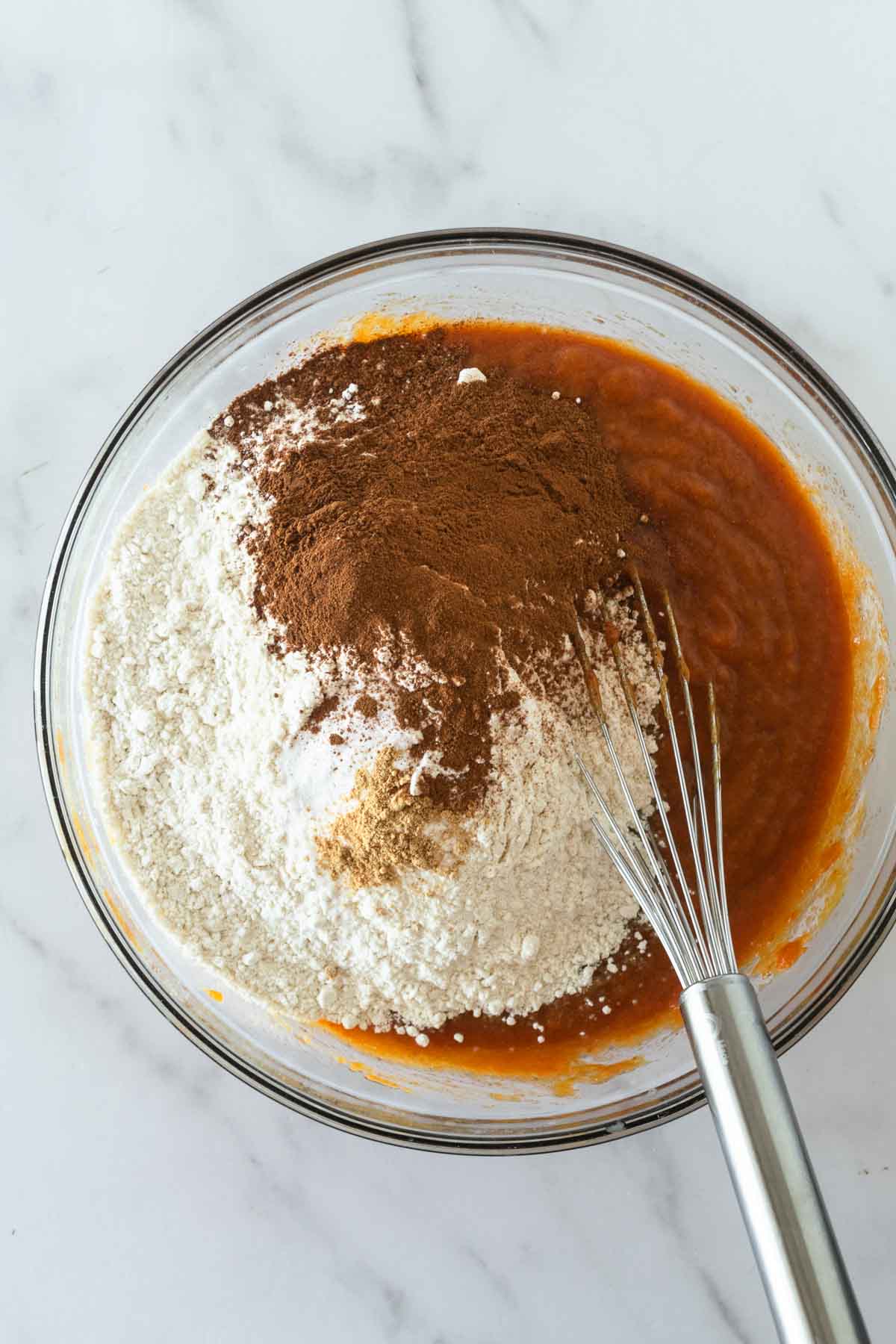 flour and spices on top of wet ingredients in a clear bowl with a whisk on a white table.