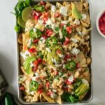 sheet pan nachos with toppings like guacamole, sour cream, tomatoes, and sour cream on a white table.