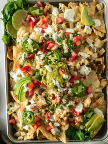 sheet pan nachos with toppings like guacamole, sour cream, tomatoes, and sour cream on a white table.