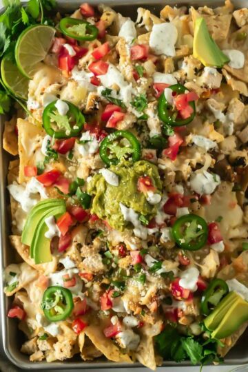 sheet pan nachos with toppings like guacamole, sour cream, tomatoes, and sour cream on a white table.