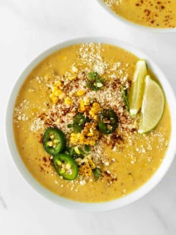 mexican street corn soup in a white bowl topped with corn, cheese, lime edges and jalapeños.