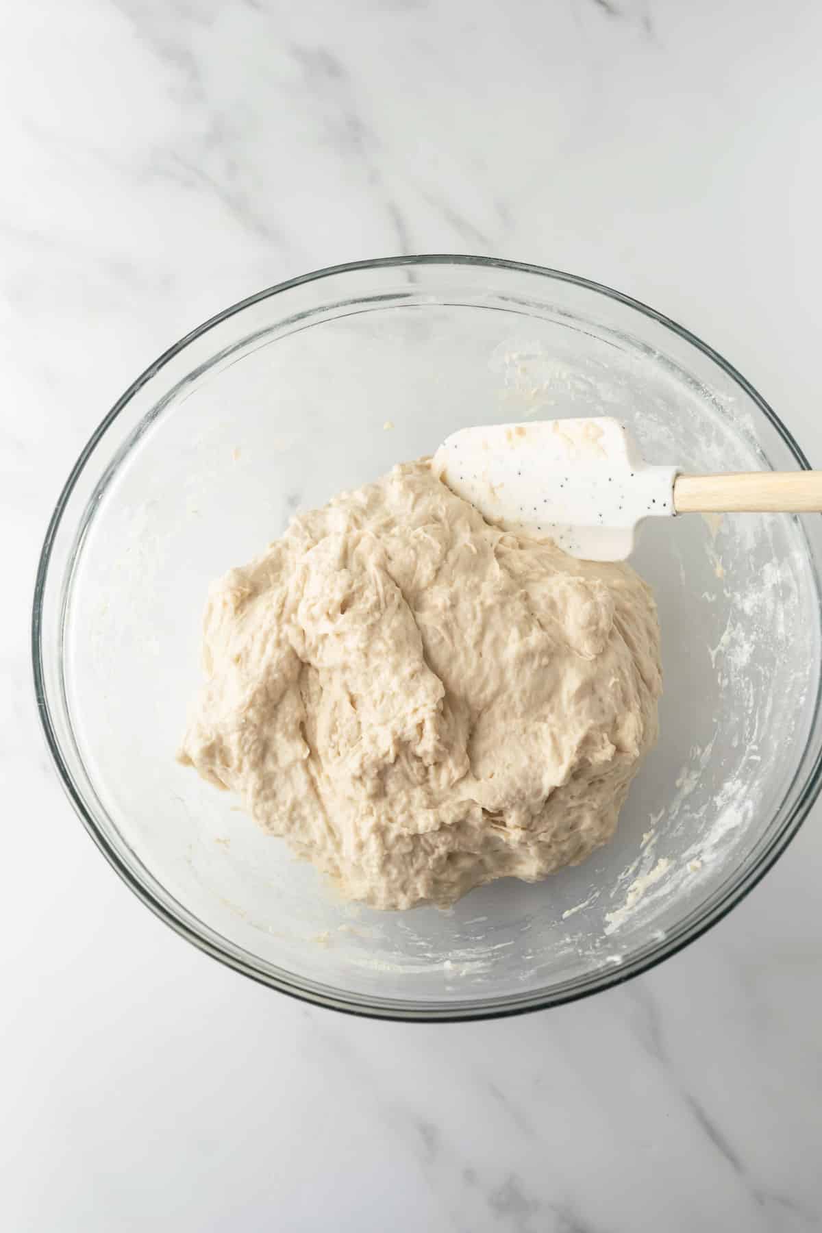 bread dough in a glass mixing bowl with a spatula.