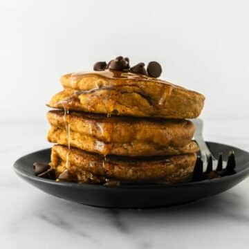 three pumpkin pancakes stacked on a black plate with a fork.