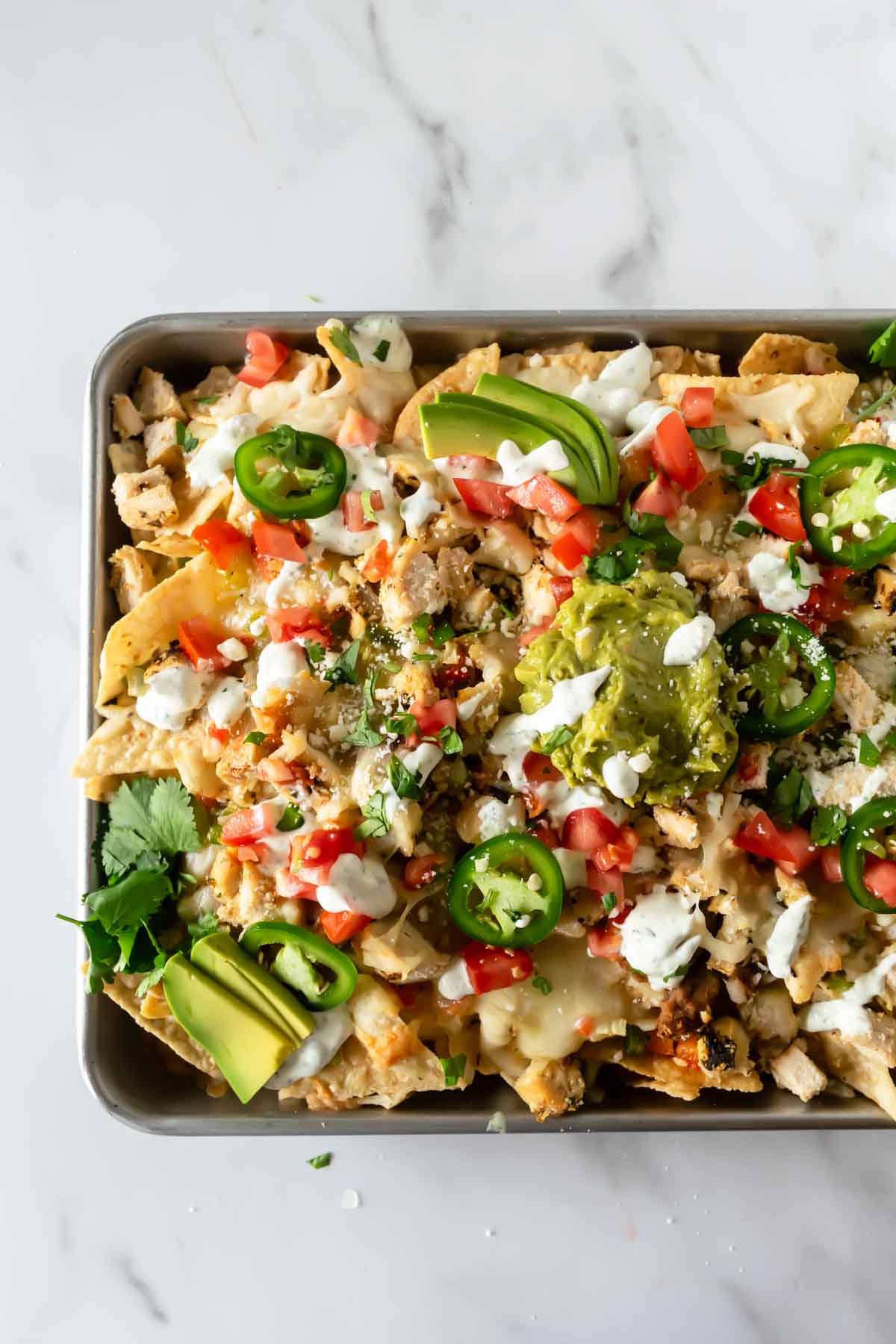 loaded sheet pan nachos on a baking sheet topped with melted cheese, tomatoes, guacamole, and jalapeños.