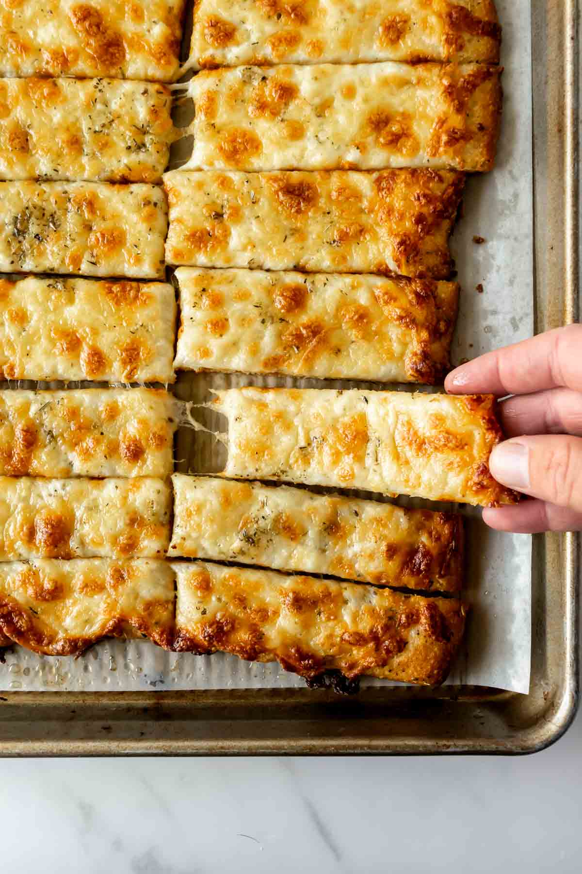 a hand reaching for cheesy breadsticks on a baking sheet.