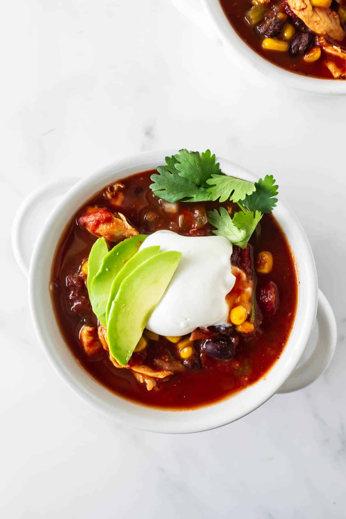 red chicken chili topped with sour cream, avocado and cilantro in a white bowl.