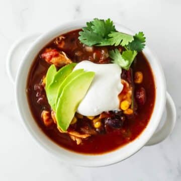 a bowl of red chicken chili topped with sour cream, avocado and cilantro.