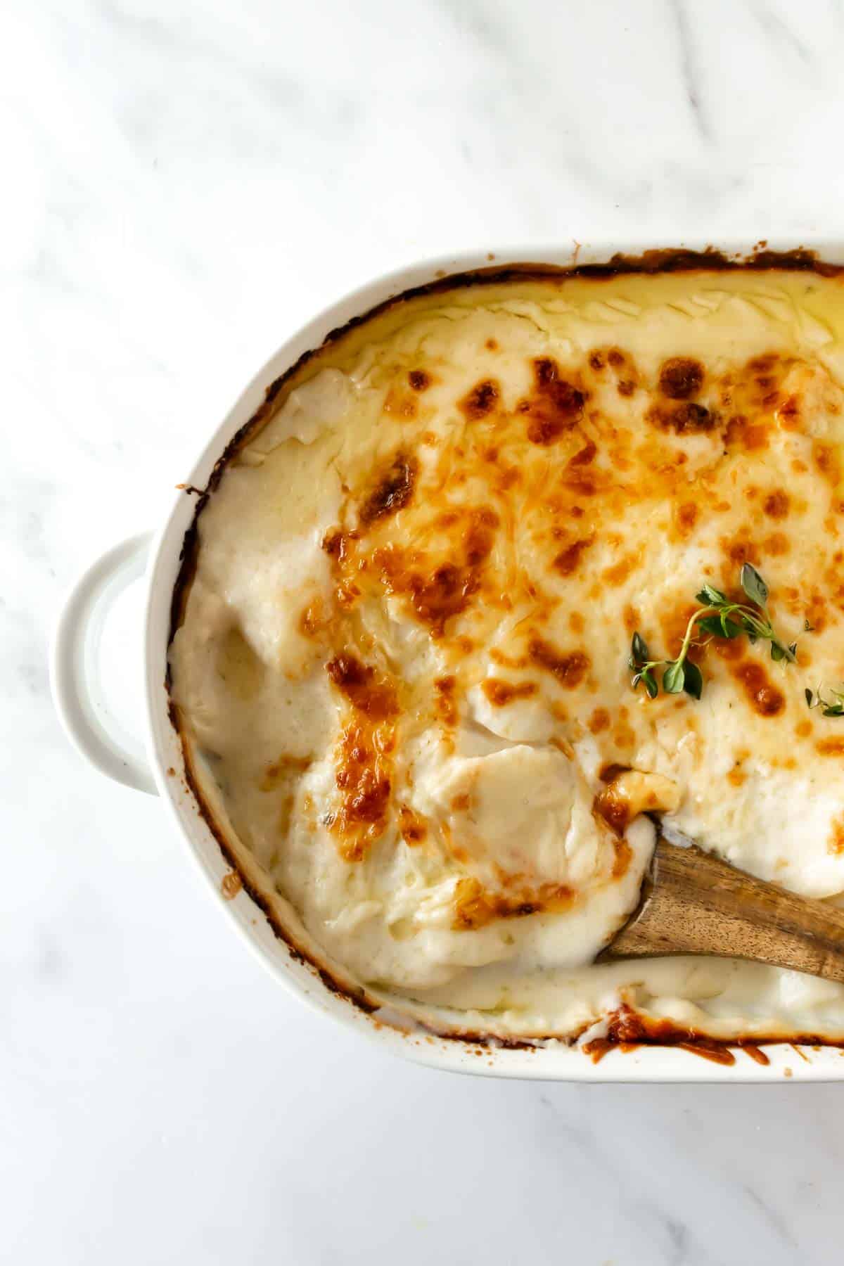 cheesy scalloped potatoes in a baking dish with a wooden spoon garnished with thyme.