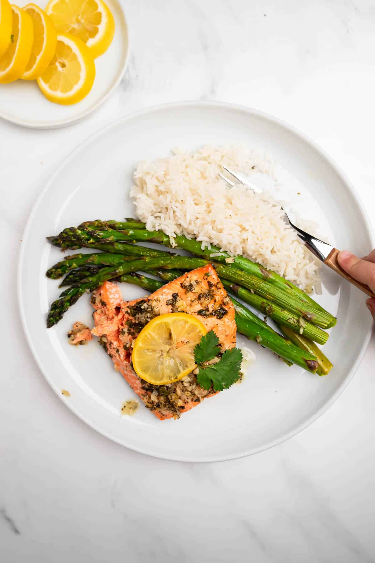 lemon garlic salmon topped with lemon slice on a white plate with rice and asparagus and a fork.