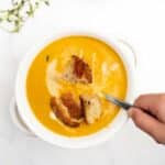 creamy pumpkin soup in a white bowl with a hand holding a spoon.