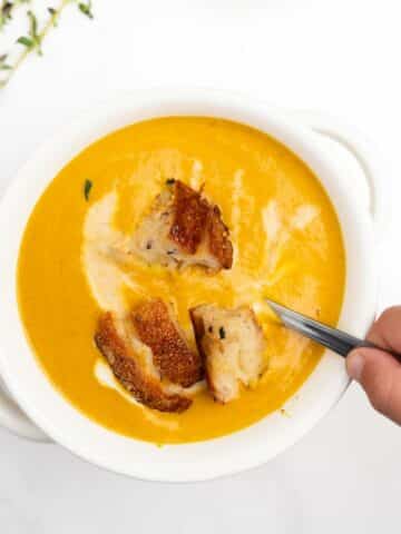 creamy pumpkin soup in a white bowl with a hand holding a spoon.